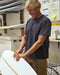 Man wearing a black short sleeve t-shirt holding a surfboard in a workshop
