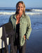 A woman wearing a green blanket shirt on the beach with ocean waves in the background